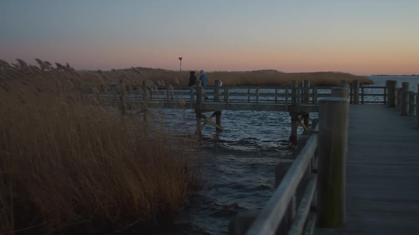 An elderly man and a young caretaker are walking on a pier on a lake early in the morning on a windy day