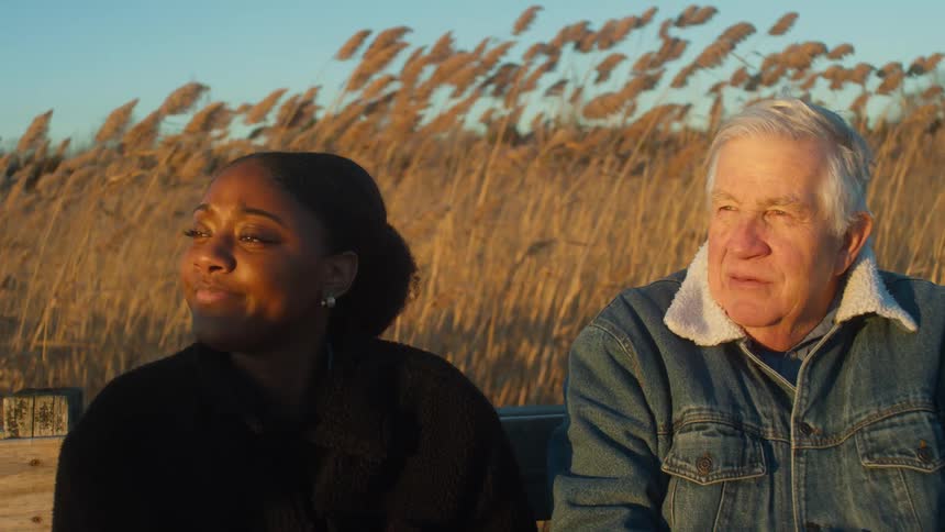 Elderly man with denim jacket and woman with bun hair and black jacket sit on bench talking to each other
