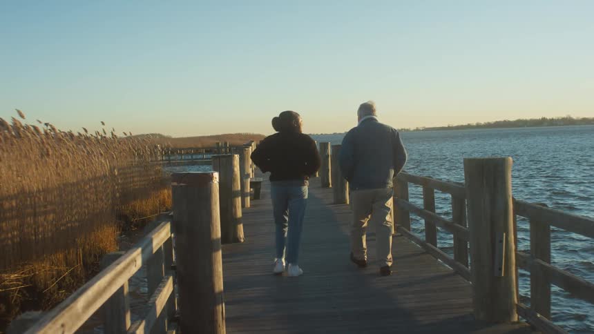 A man and woman are walking outdoors on a wooden pier by a large lake in a rural location at sunset