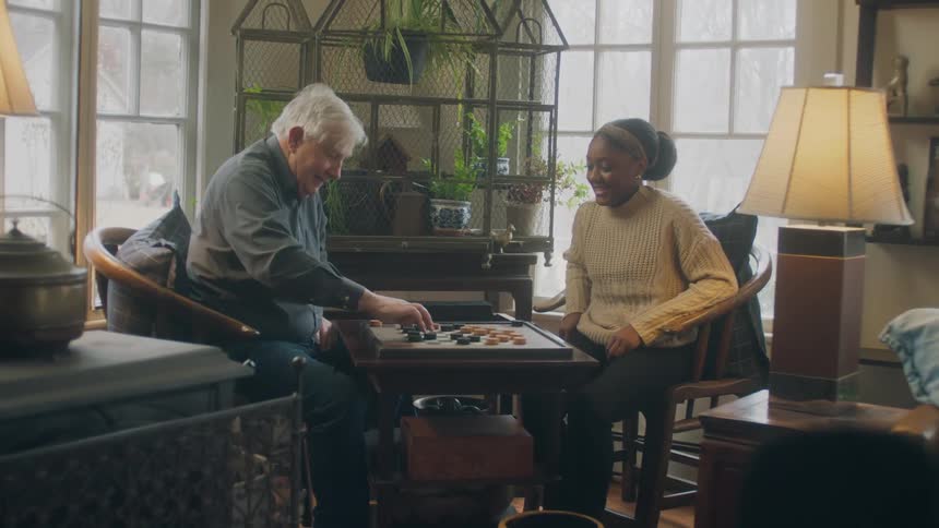 An elderly man makes a move on the checkerboard while a young woman smiles