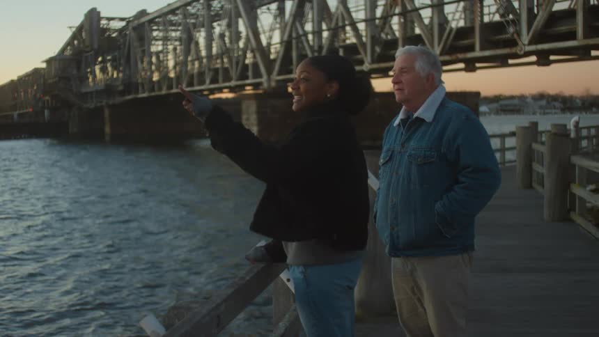 A young woman and an elderly man stand on a dock and talk as they look out at the water