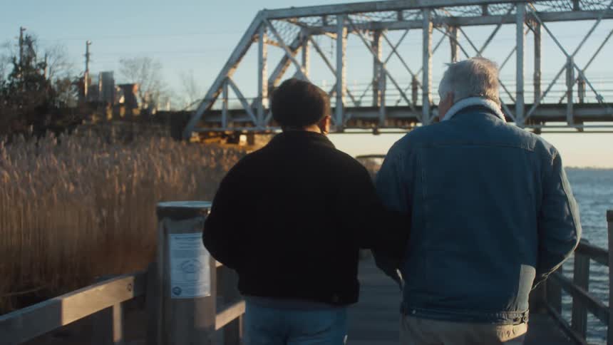 Elderly man and young woman walk on the pier near the lake as a bridge crosses the lake in front of them