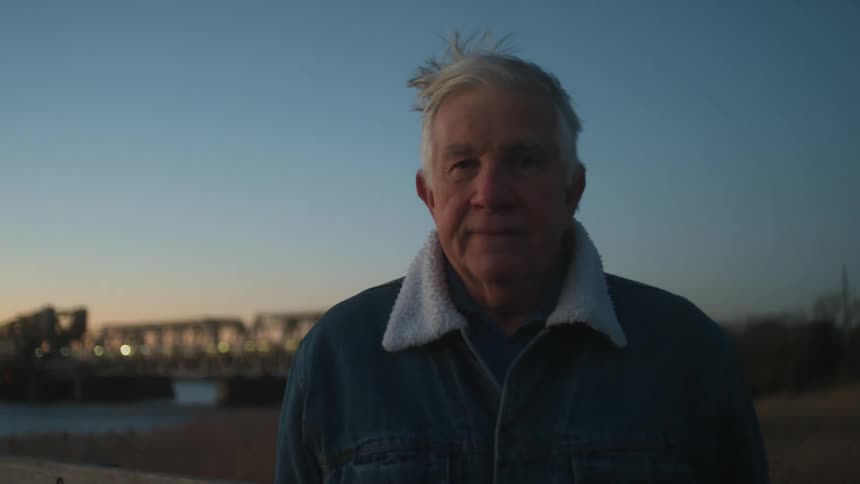 An elderly man is standing on a boardwalk and staring at the camera at dusk with a passenger train passing in the background