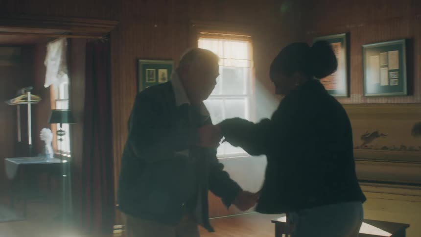 Elderly man with denim jacket and jeans dances holds woman and and dances together next to piano in room