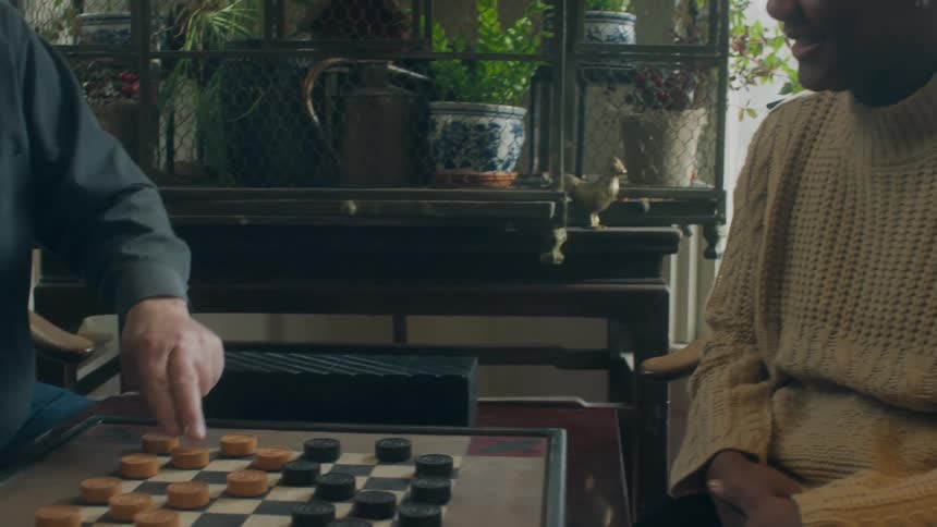 Elderly man and woman play checkers sitting in room near plants sitting on table behind a mesh enclosure