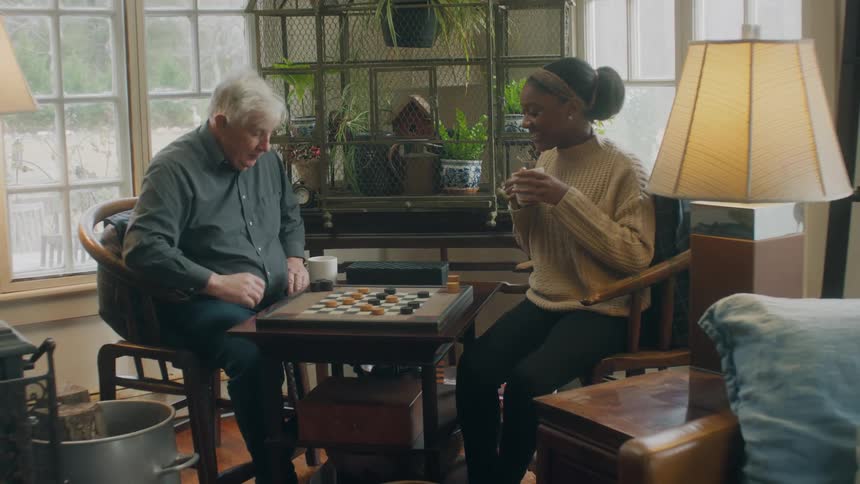 An elderly man and younger woman drinking coffee and playing checkers