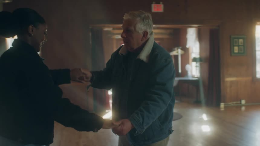 An elderly man and young woman dance together in an open hall