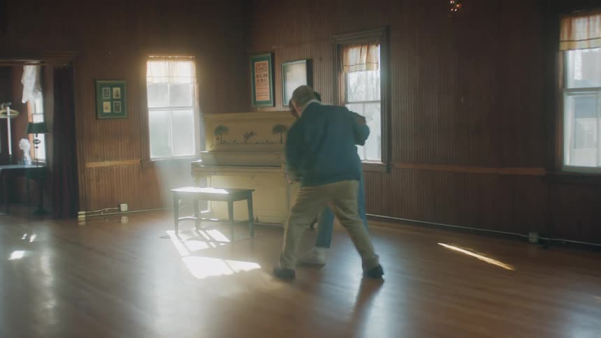 Old white man and young black woman dancing together in the middle of an empty room next to a white piano
