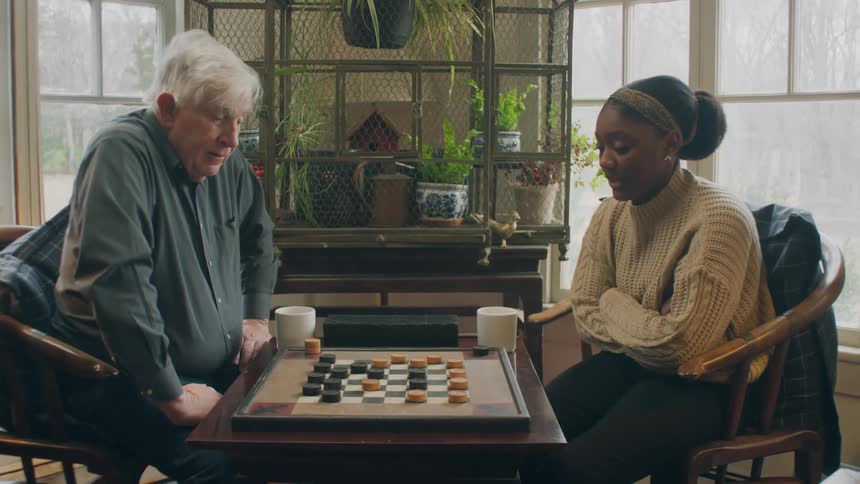 Elderly man with dark shirt and jeans sits in armchair with woman with bun hair and white knitted sweater playing checkers in room