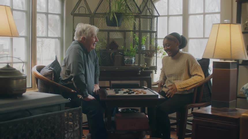 Old white man and black woman sitting indoors next to the windows playing checkers and having fun together