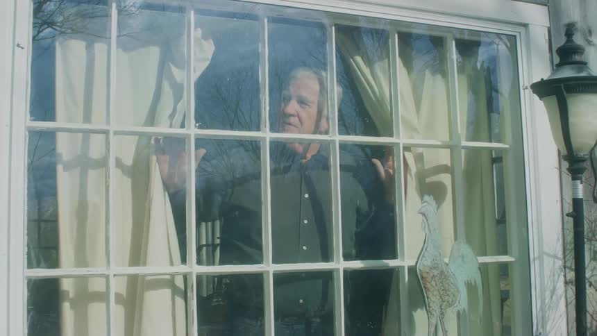 Elderly man with dark shirt and jeans opens the curtains and looks out the window in the morning