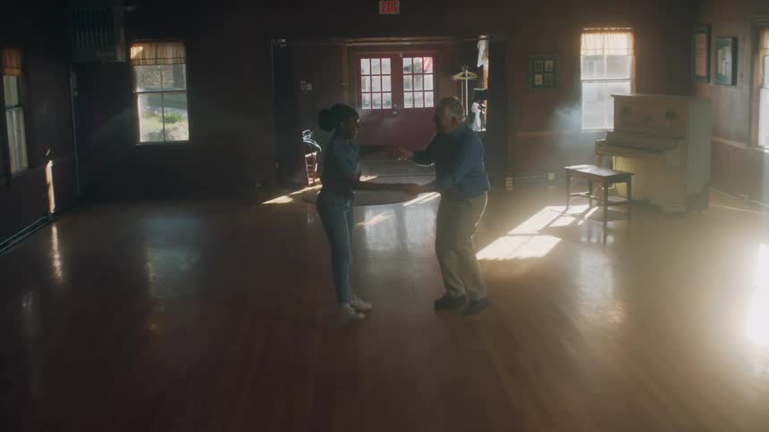 Elderly man and young woman hold hands as they dance across a large room as a piano sits in the corner