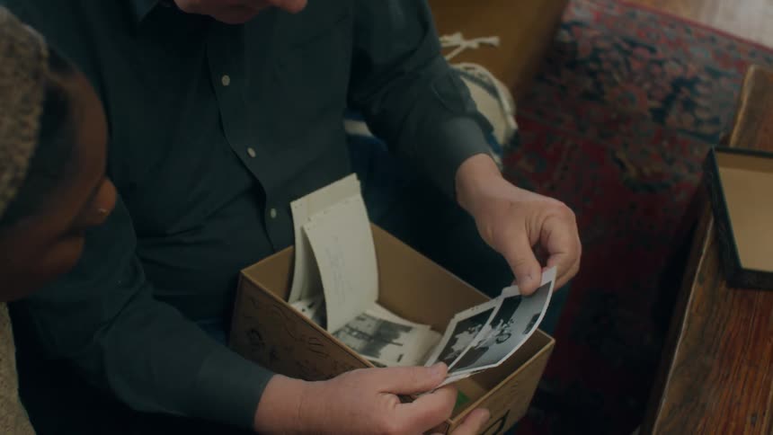 A young woman sitting next to an elderly man as he goes through photographs in a box and tells her about them