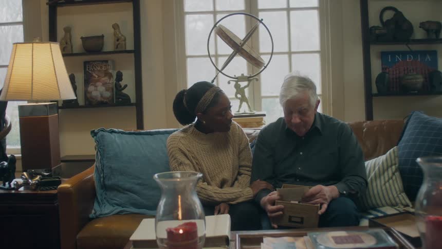 A young woman sits next to an elderly man and comforts him as he reads an old paper from a box