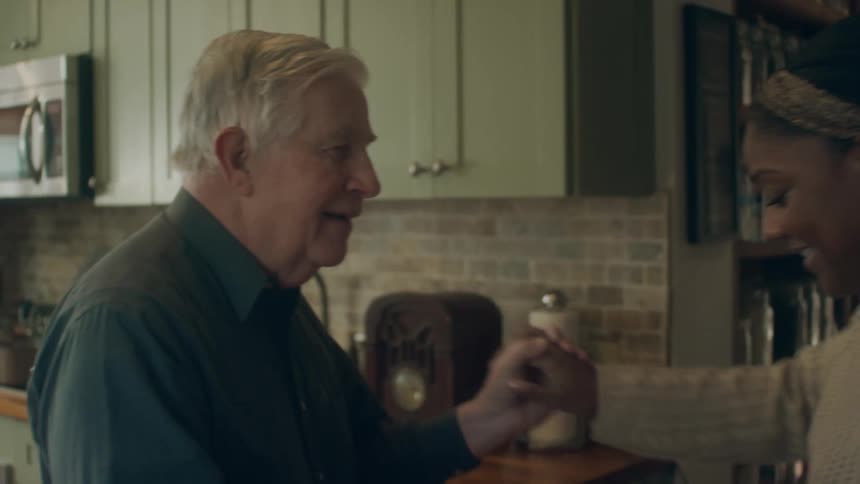Woman with white knitted sweater dances with elderly man wearing dark shirt in kitchen in the living room