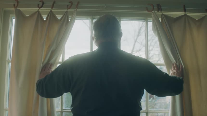 An elderly man pulling back curtains and looking out the window
