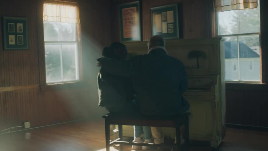 Older man sitting at piano welcomes woman with a hug when she walks away from window to join him