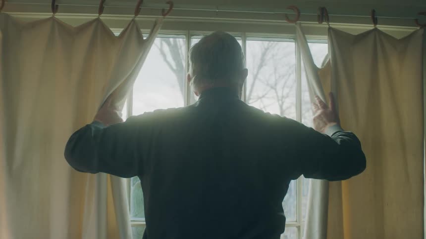 Old white man opening the window curtains and looking outside through the window