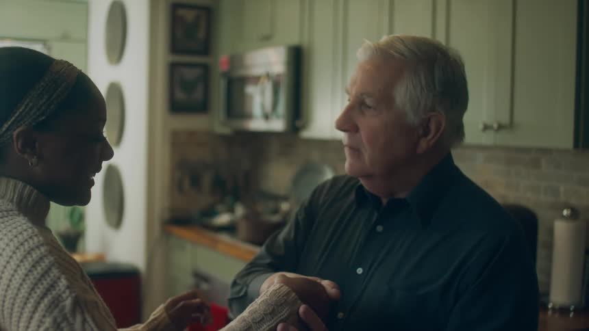 Old white man inviting and dancing with a black woman inside the house while there's an old radio playing above the kitchen cabinet