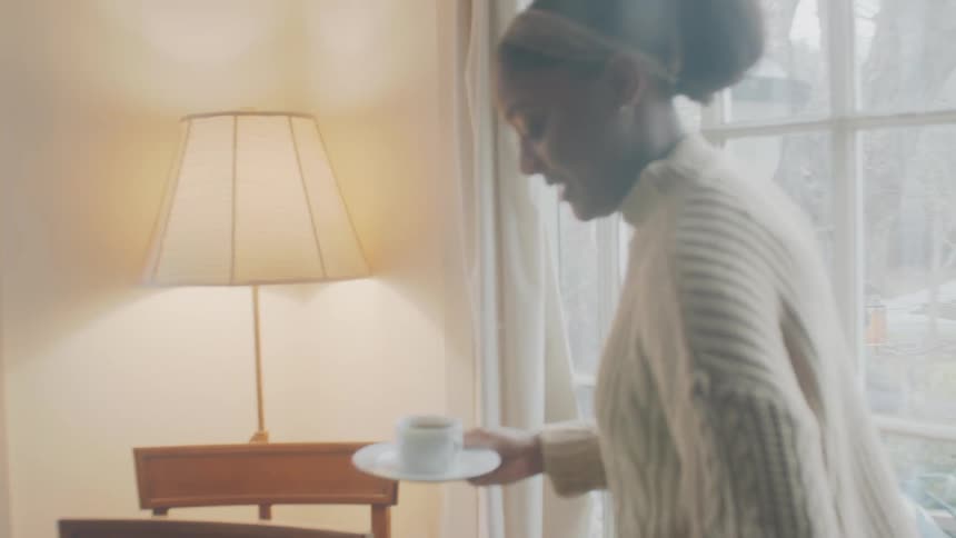 An elderly man and a young woman walk down a hall in a home carrying mugs of coffee and set them on a table by a laptop