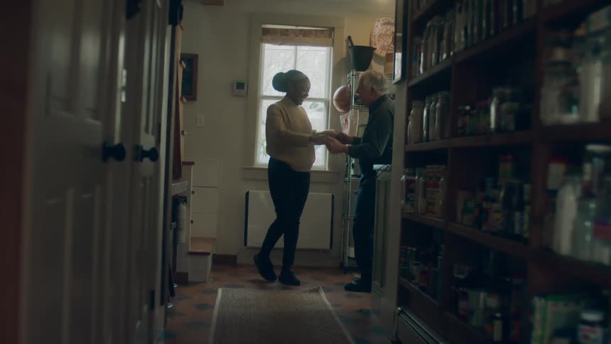 A man and woman dance in their kitchen at the end of a long hallway