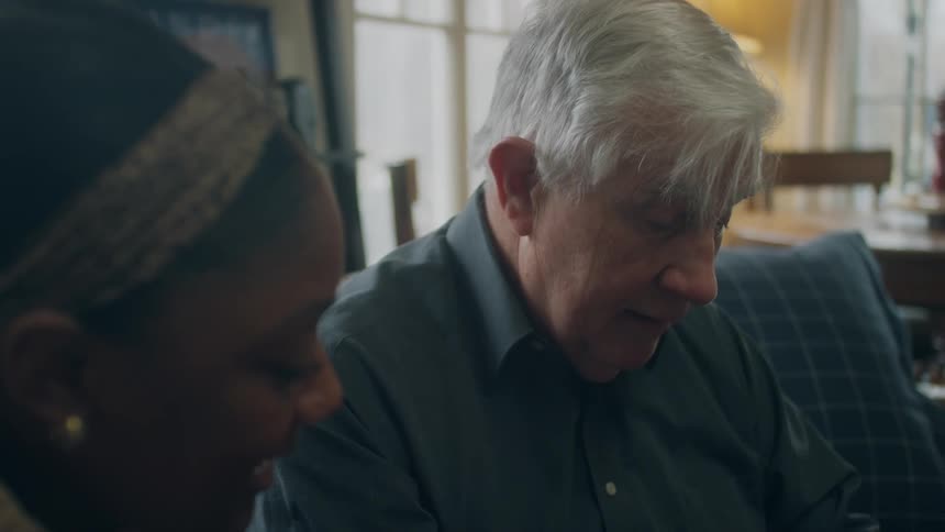An elderly man sits at home next to a young woman and they talk and laugh