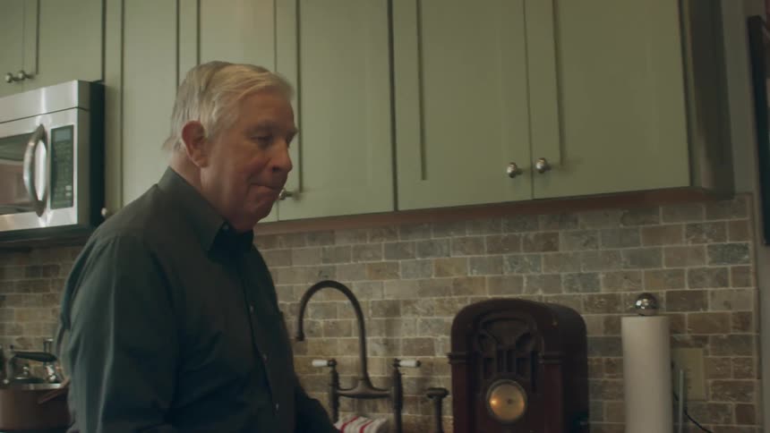 Old white man turning on an old radio at the kitchen cabinet and inviting someone else with his hands to dance with him