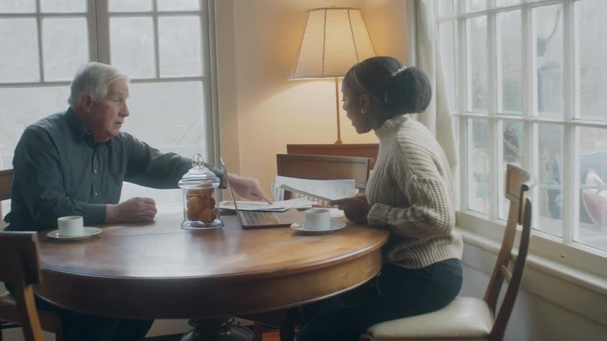 Elderly man points to some papers as he and a young woman sit at a table with a laptop