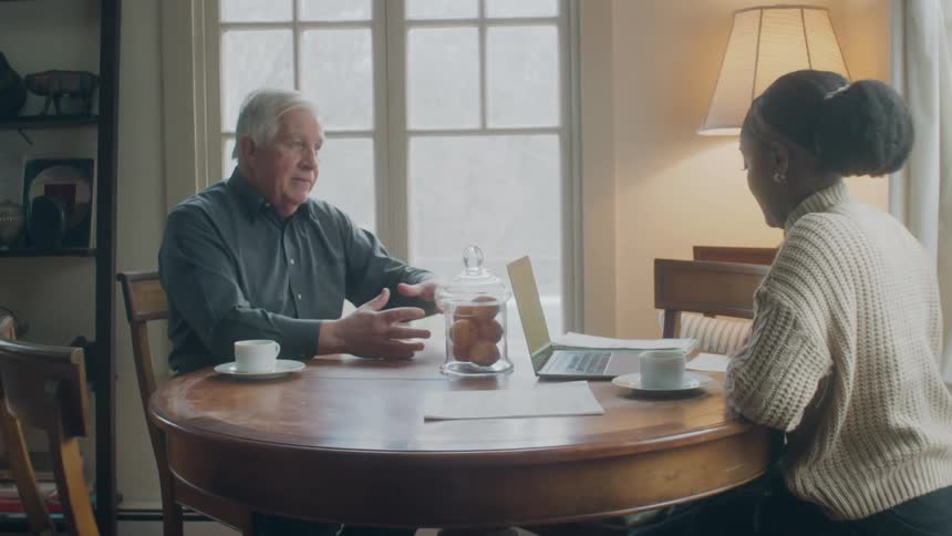 A young woman sits a table with a laptop and a cup of coffee and talks with an elderly man