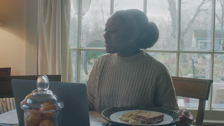 Woman with bun and white knitted sweater sits at table with food talking to elderly man wearing blue shirt at home