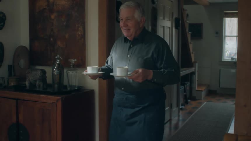 Man walks into room carrying coffee cups and serves one to a woman sitting at table in front of a laptop