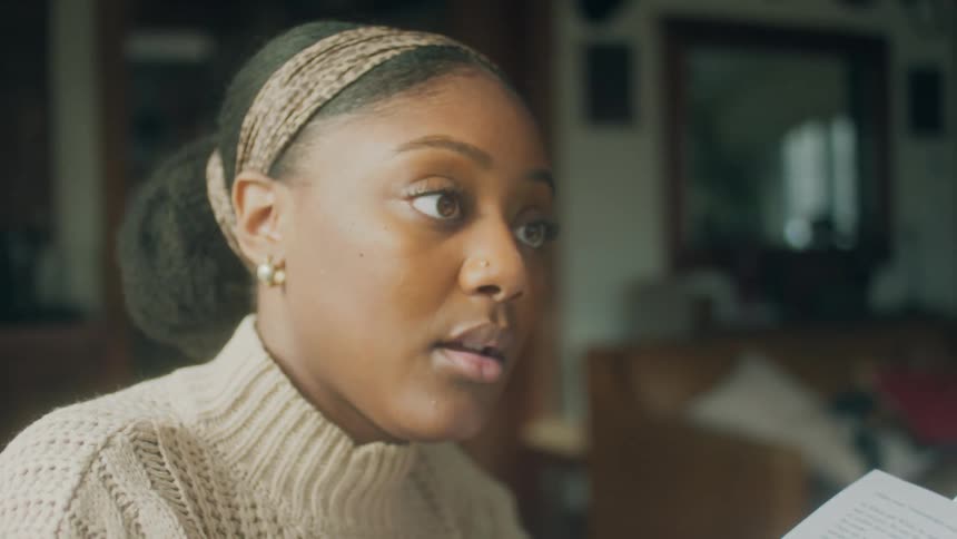 Black woman sitting in the living room reading a white paper and talking to someone else