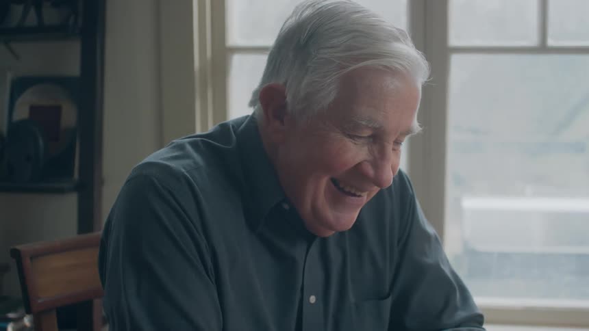 A man laughs as he sits at a table inside his home