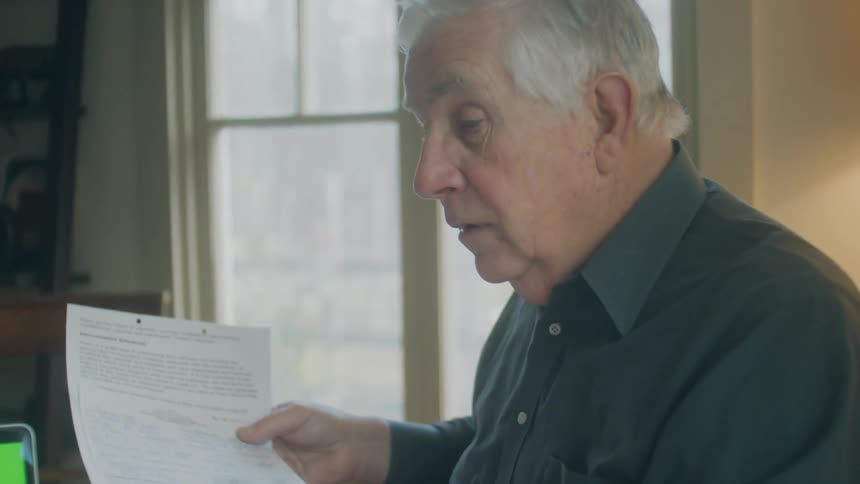 An elderly man at home holds a document and talks with a woman in front of a laptop