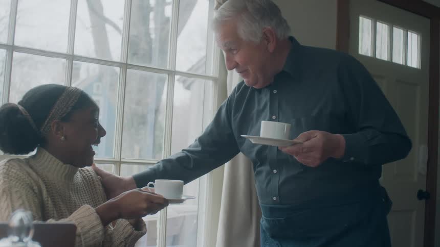 Elderly man carries two cups and saucers and hands one cup and saucer to a woman sitting in a chair