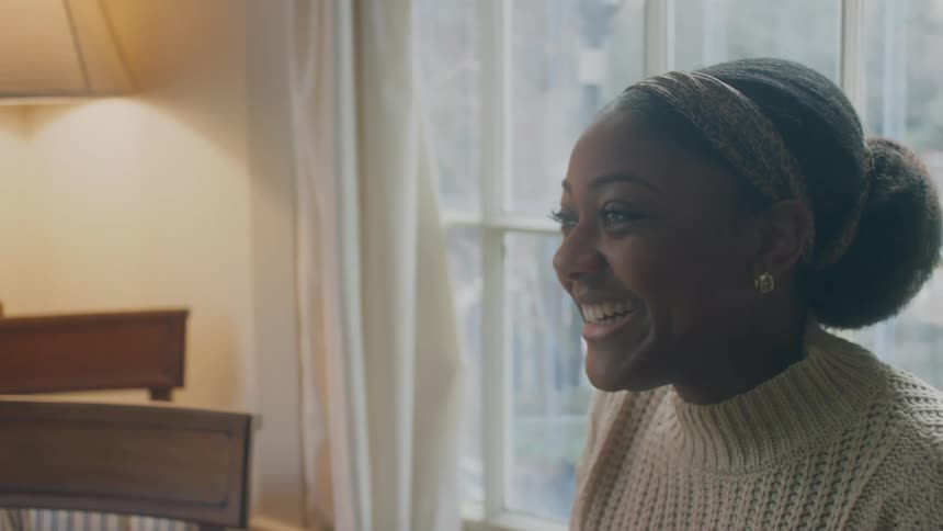 A woman smiling and laughing as she talks sitting at home in front of a window