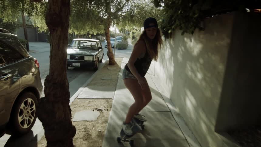 A girl skateboards on the sidewalk of a car lined street