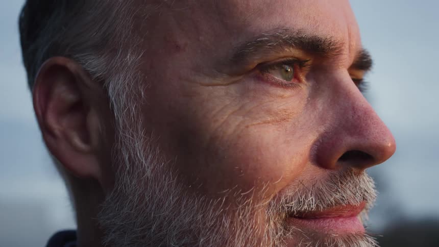 White old man with green eyes and a beard standing outdoors staring ahead and smiling when the sun light reaches his face