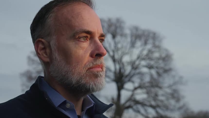 A man with a gray beard is standing outside with a concerned look on his face and there is a bare tree behind him