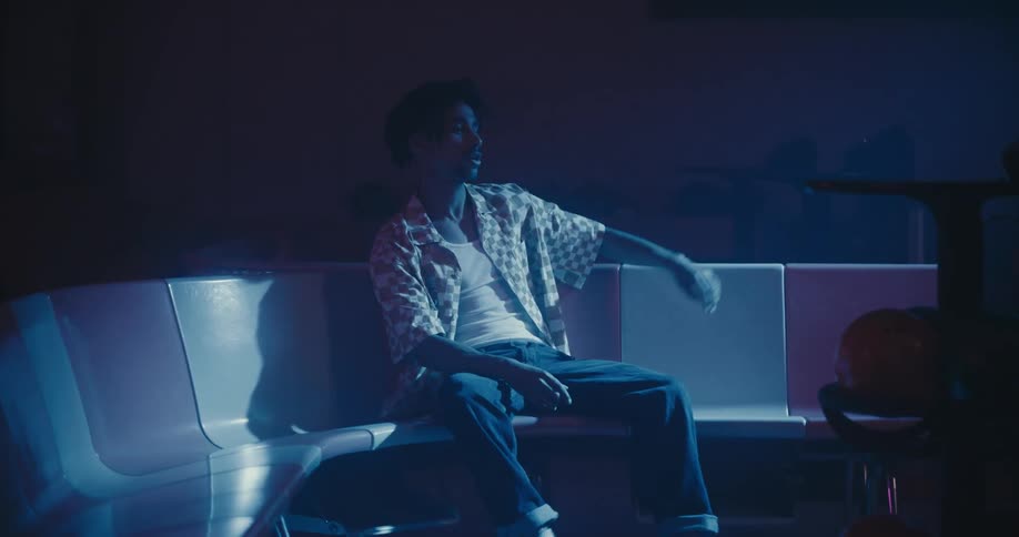 A woman gets up from bench at a bowling alley while her boyfriend stays and bobs his head to music