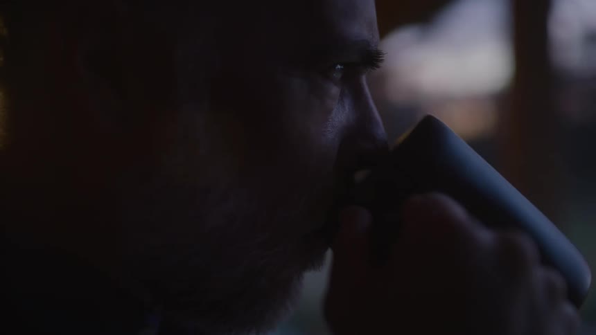 Old man standing at the front porch of his house looking ahead and drinking from a cup