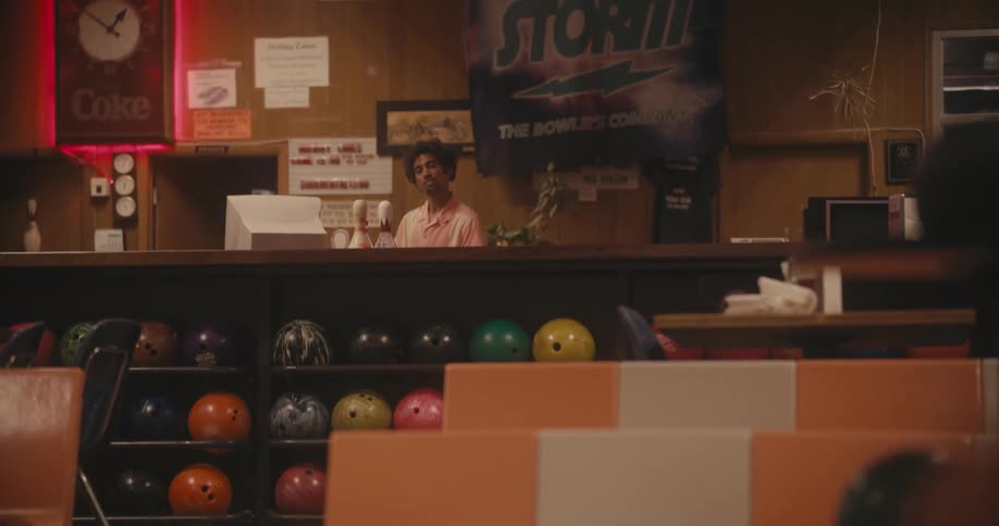 Man stands at bowling alley counter while a woman picks up a bowling ball in a lane