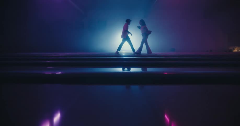Man and woman dance on walkways above glossy reflective surface in dark area lit by bright spotlight