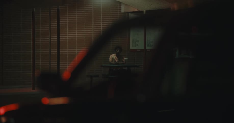 Man is visible through parked cars side window and windshield as he sits at table under flickering light of building