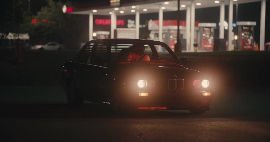 A man sits in a parked car near a gas station as traffic passes by