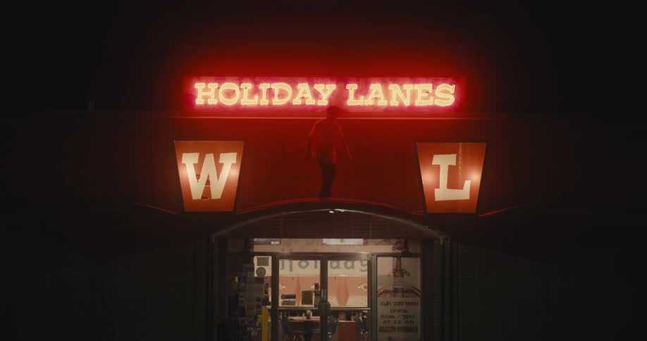 Young man dancing in front of the neon sign on the roof at the entrance of a bowling alley at night
