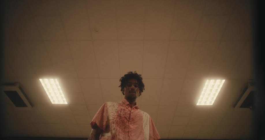 Black man breathes heavily while looking down as two ceiling lights illuminate the room