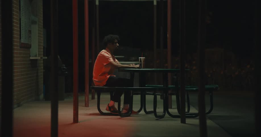 A man sitting alone at an outdoor table at a restaurant near the parking lot with a light flickering