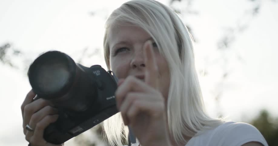 A blond woman gestures while holding a camera