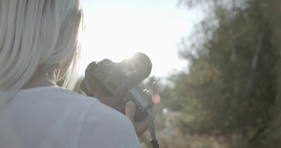 A blond woman holds a camera and looks toward the sun
